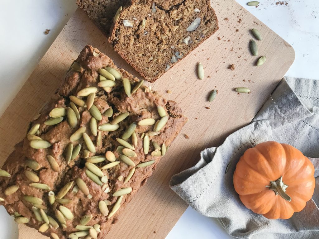 A loaf of Spiced pumpkin, maple and pecan bread laid on a wooden board with two slices cut. The loaf is topped with pumpkin seeds.